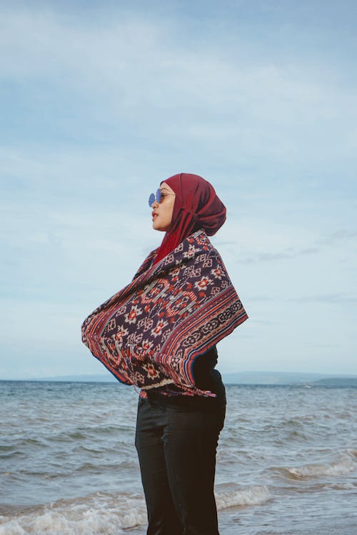 Low angle side view of tranquil Muslim woman wrapped in scarf enjoying solitude near waving sea