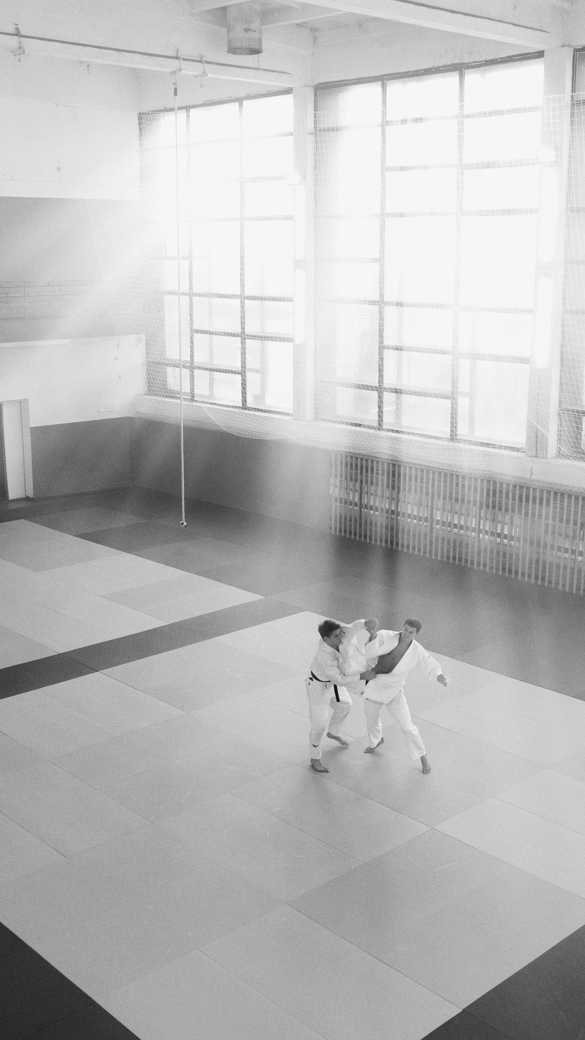 Two martial artists train in a sunlit dojo, captured in black and white.