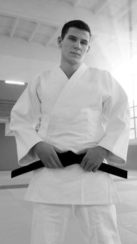Black and white portrait of a martial artist wearing a gi, holding a black belt indoors.
