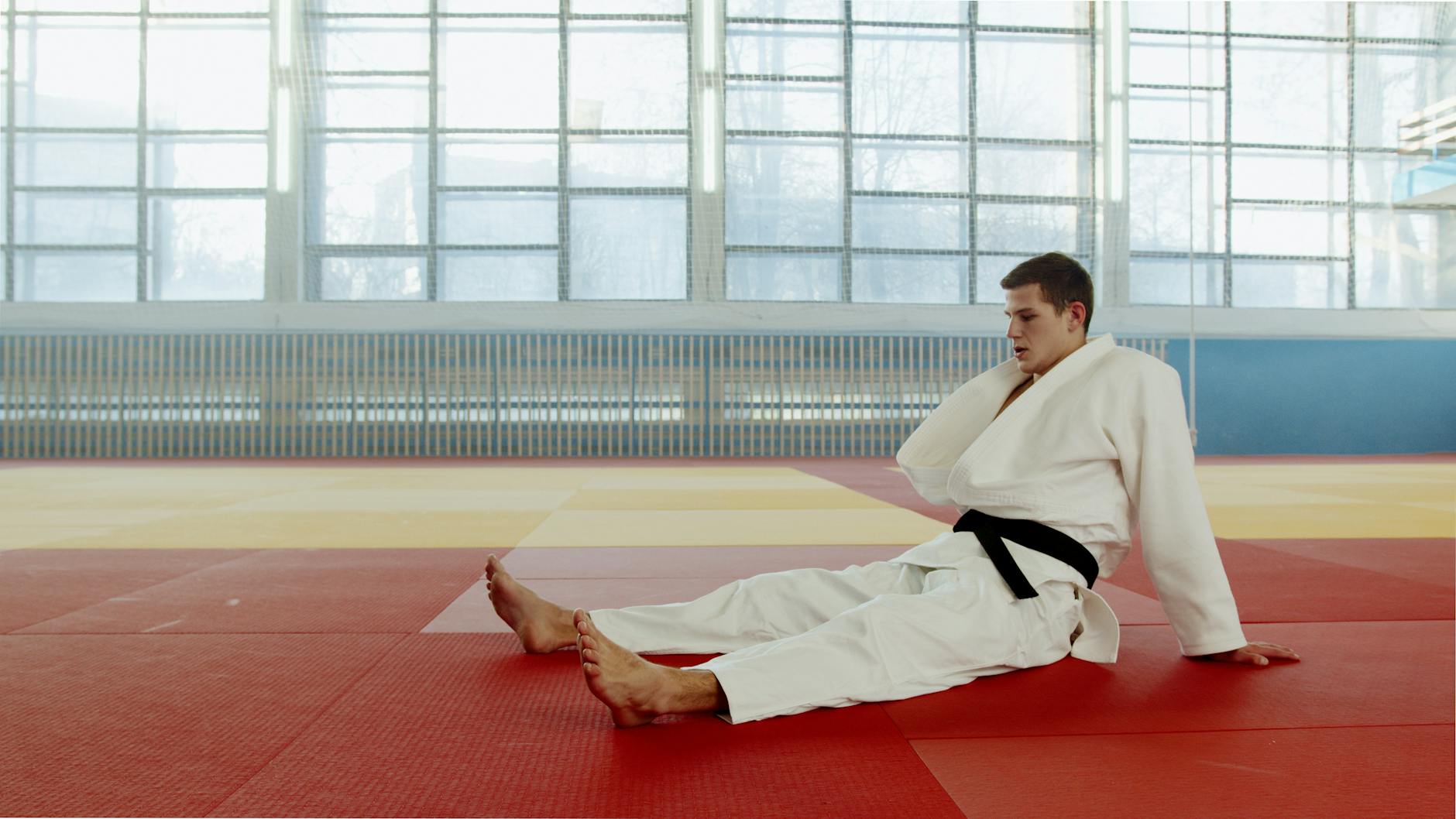 Sharpens Mental Focus & Builds Resilience 2 A martial artist in a white uniform with a black belt rests on an indoor gym floor, looking contemplative.