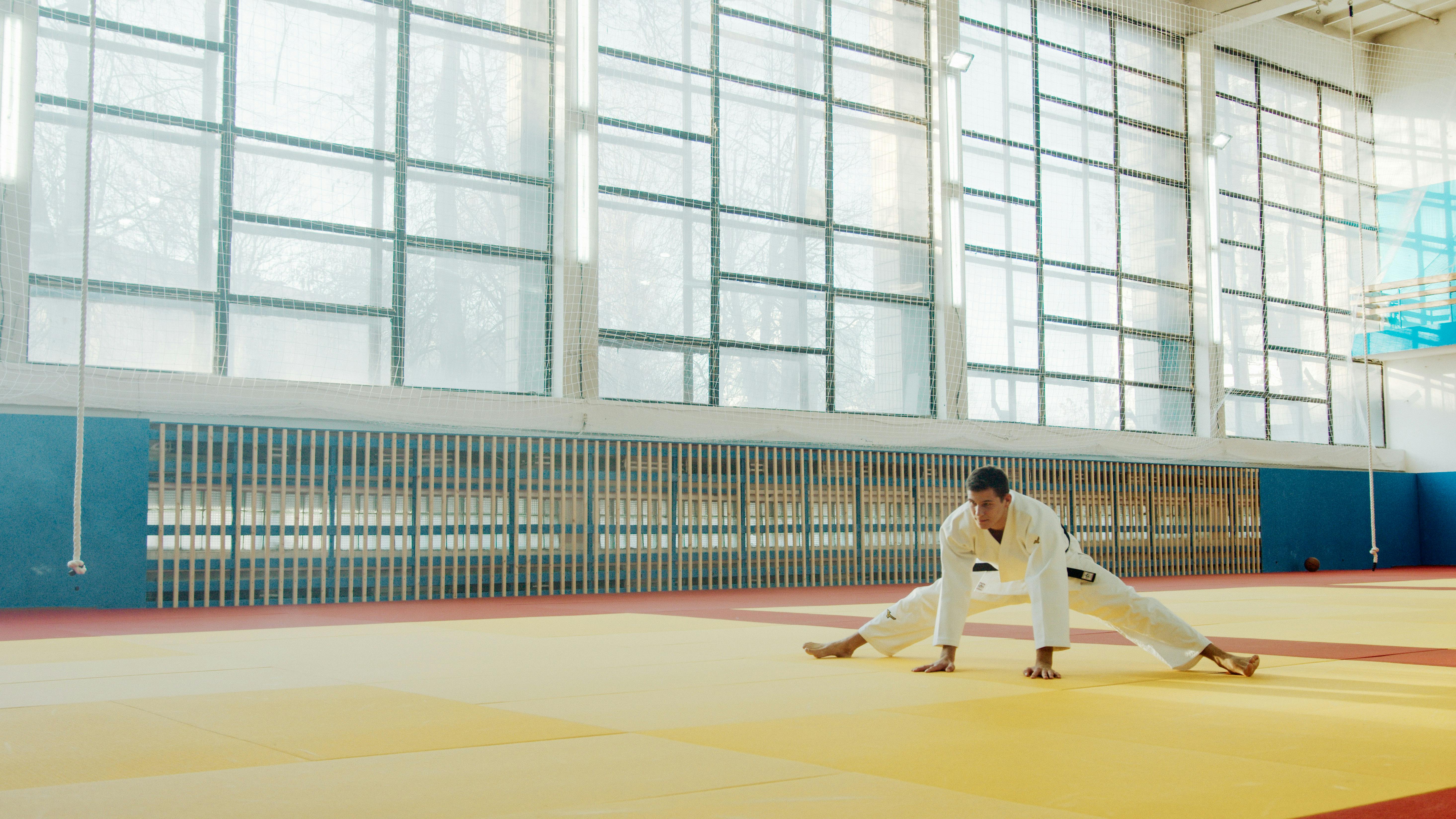 Martial artist in uniform stretching on a training mat in a bright indoor gym.