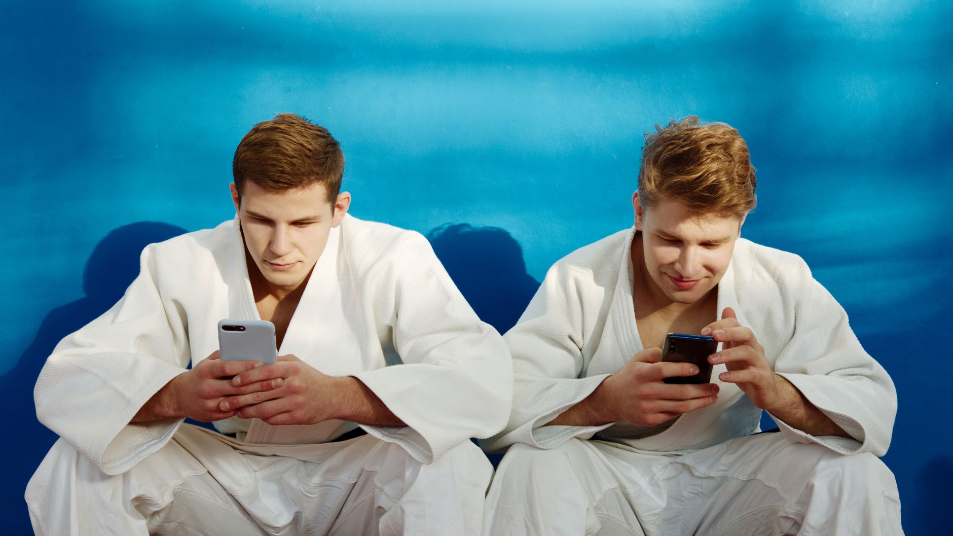Two martial artists in gis using smartphones against a blue background.