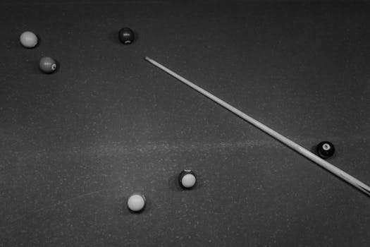 Artistic black and white photo of a billiards table with scattered balls and a cue stick.