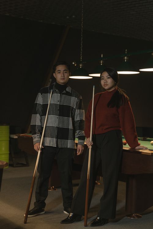 Free A young Asian couple enjoys playing billiards indoors, posing with cue sticks under dim pool room lighting. Stock Photo
