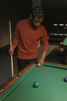 A man in a red sweater lines up a shot in a dimly lit pool hall, holding a cue stick.