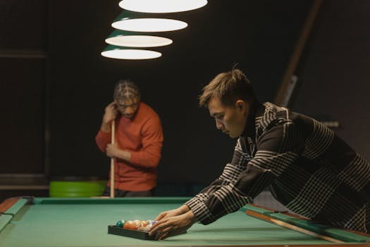 Two men setting up a billiards game indoors under warm lighting.