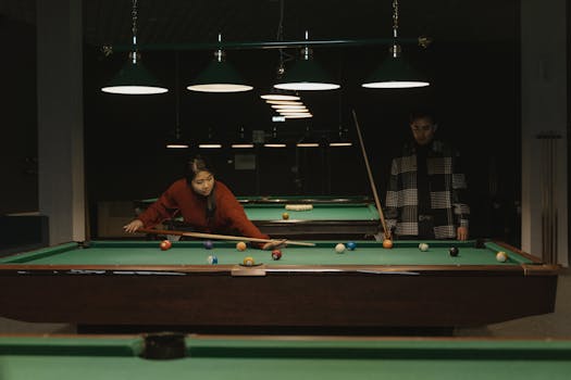 Two adults playing billiards under dim lighting, focusing on shot angles.