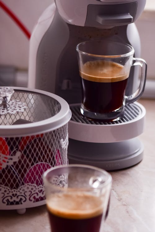 Free Modern professional coffee machine pouring fresh coffee into glass cup placed on wooden table near capsules in light kitchen at home Stock Photo