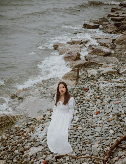 From above of gentle ethnic female tourist in white dress looking at camera from pebble ocean shore