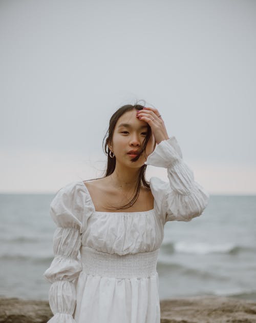 Young tender ethnic female traveler in white apparel looking at camera against wavy sea in daytime