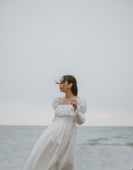 Young tender ethnic female tourist in white dress admiring wavy ocean while looking away during trip in daylight