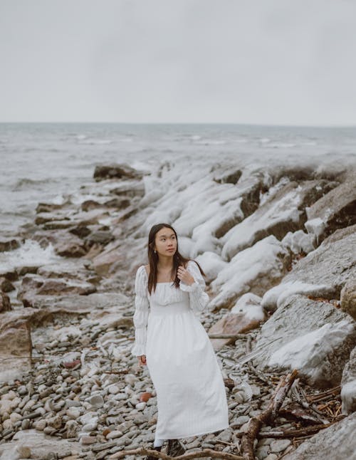 Free Young contemplative ethnic female tourist in white dress looking away on pebble beach against endless ocean Stock Photo