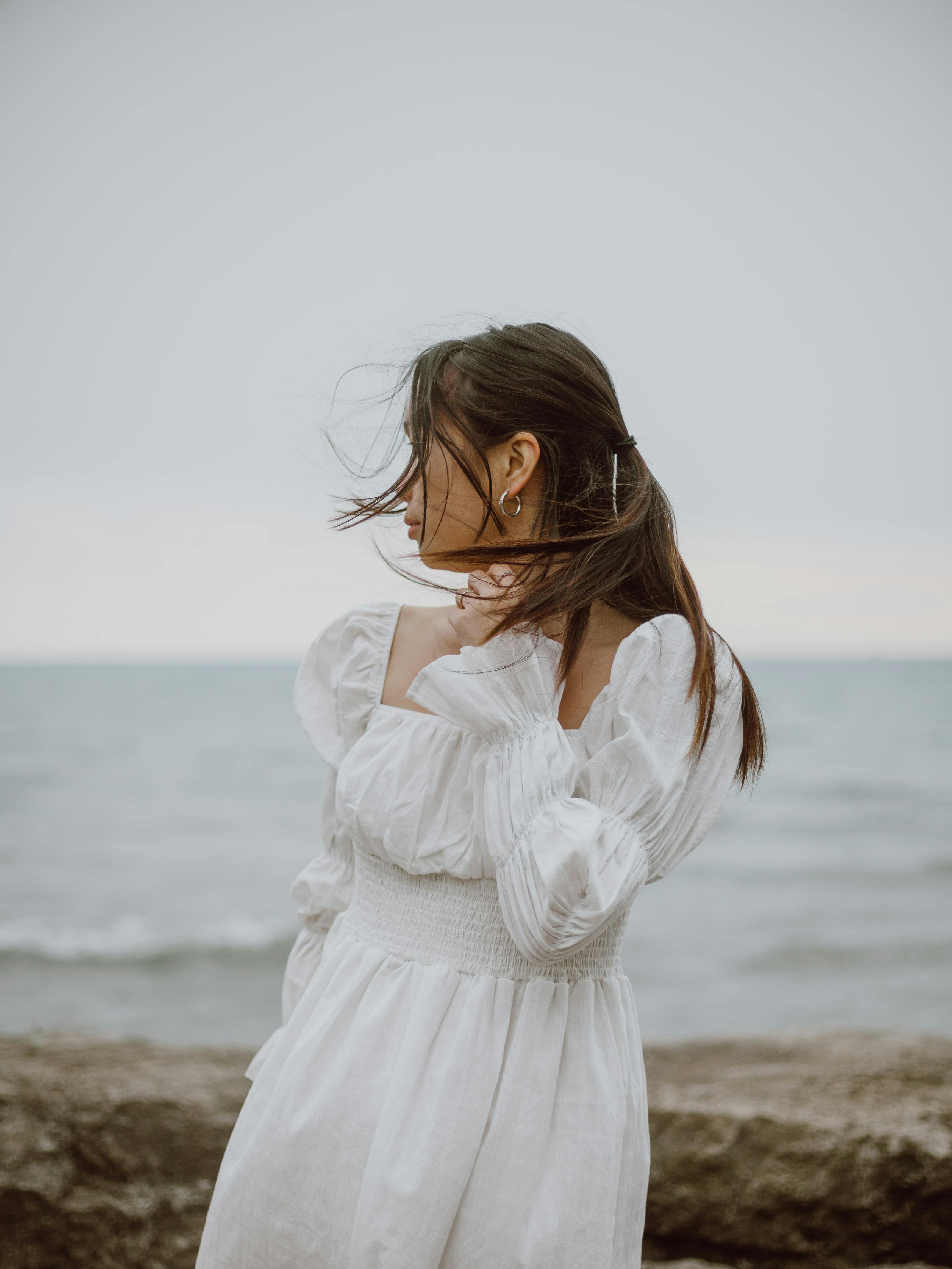 Young contemplative female traveler in dress touching neck while looking away from coast against ocean