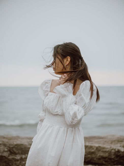 Young contemplative female traveler in dress touching neck while looking away from coast against ocean