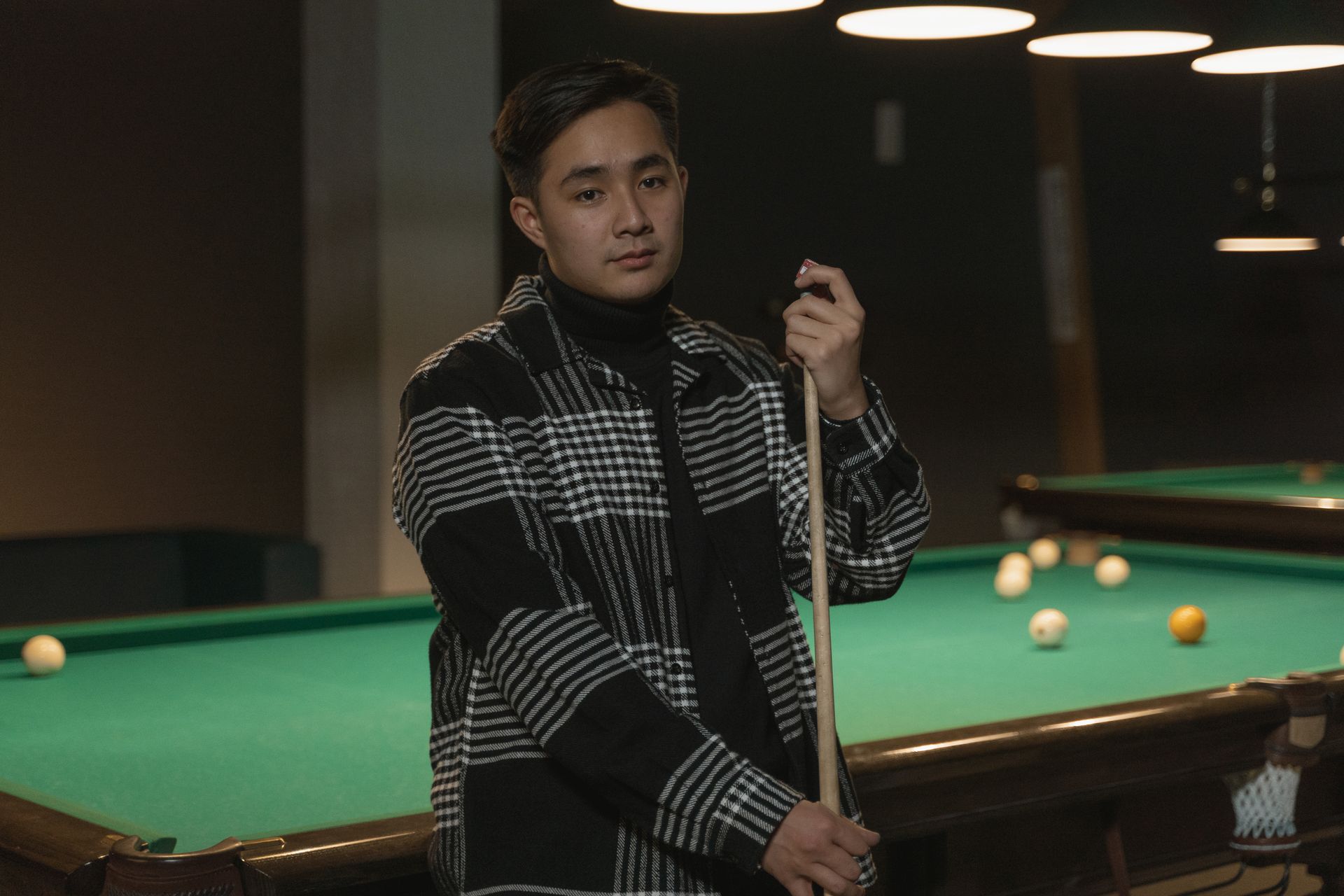 A young man stands confidently holding a cue stick beside a billiard table in a dimly lit pool hall.