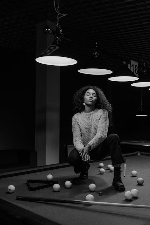 Woman in Sweater and Pants Sitting on Billiard Table while Posing at the Camera