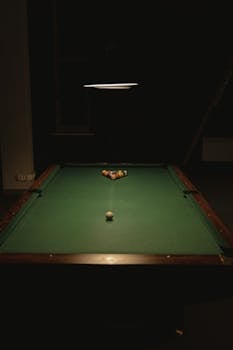A billiard table with arranged balls under a focused ceiling lamp, evoking a tense atmosphere.