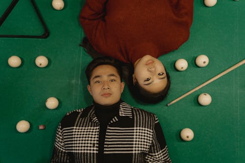 A Man and a Woman Lying on a Billiard Table Surrounded by Billiard Balls while Seriously Looking at the Camera