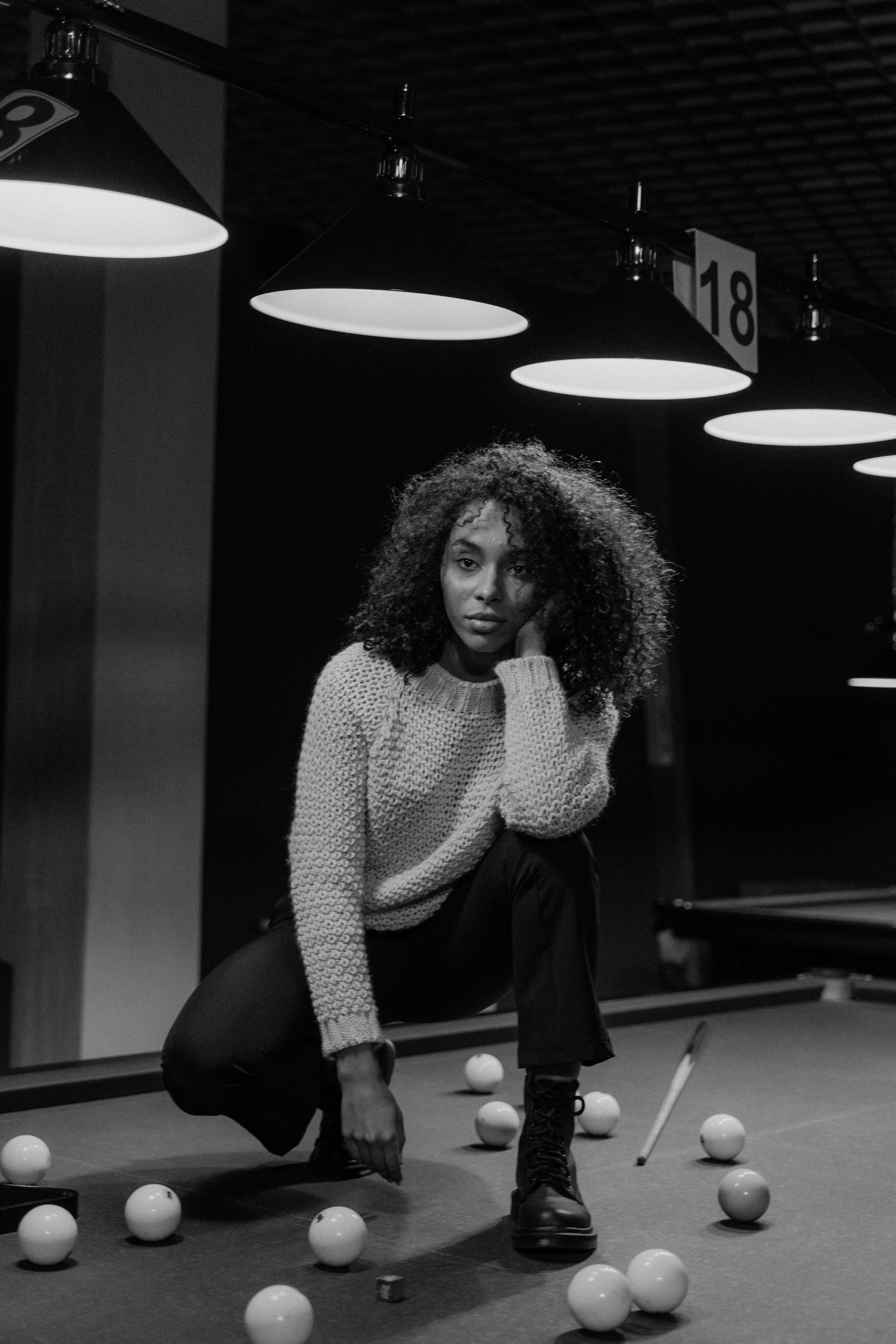 grayscale photo of a woman with curly hair posing on a pool table