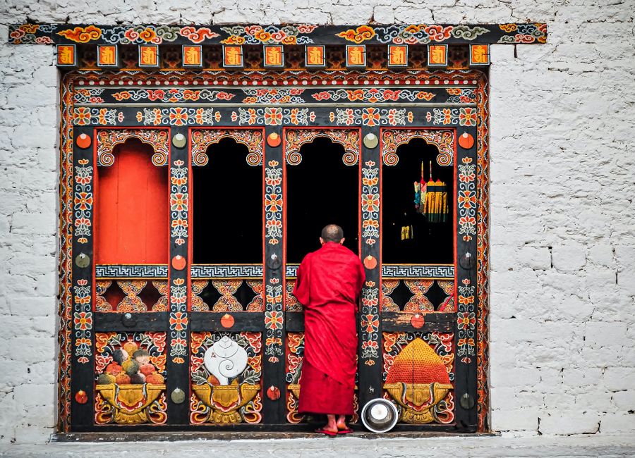 A Buddhist monk in red robes standing at an ornately decorated temple doorway in Bhutan.