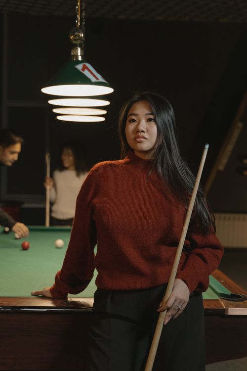 Free Confident Asian woman in a billiards room holding a cue stick with friends playing pool in the background. Stock Photo
