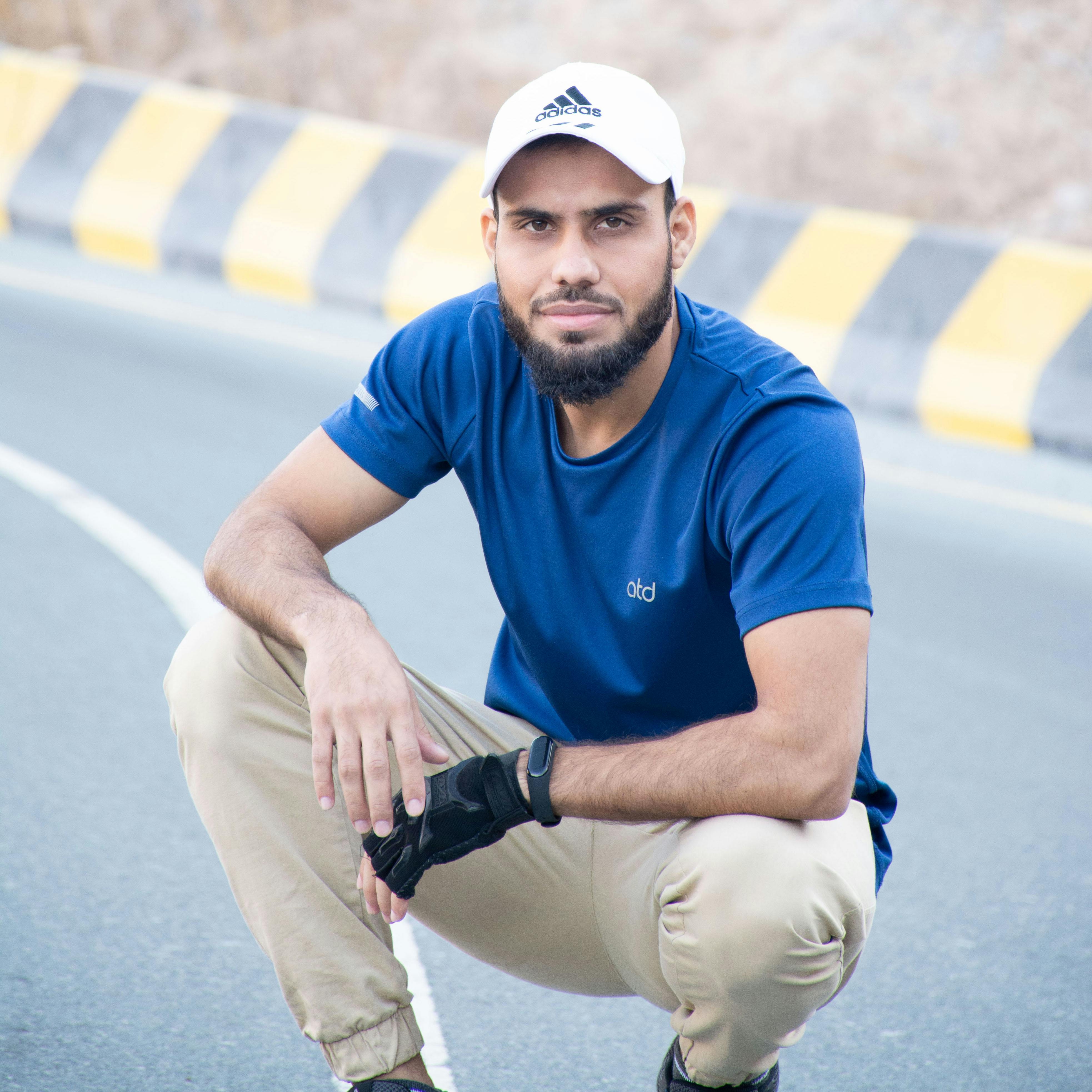 Bearded man wearing blue shirt and cap crouching on urban street.