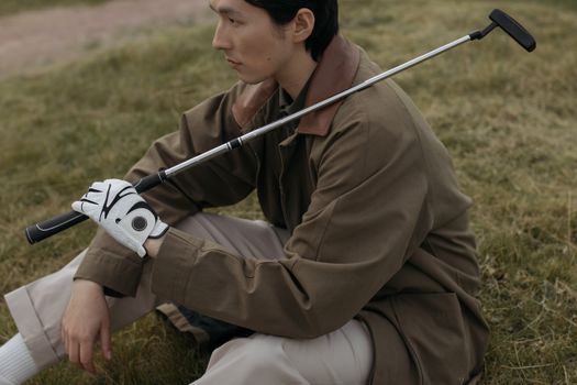 A young man in a coat sits on grass holding a golf club, reflecting a serene outdoor moment.