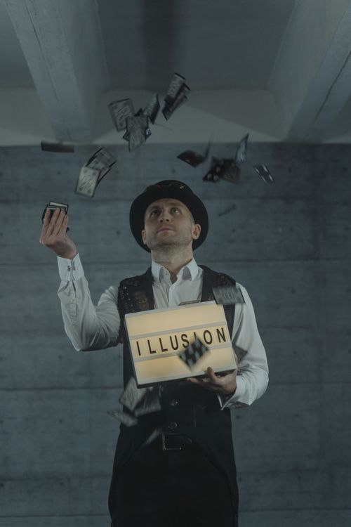 Free A magician performing a card trick with an 'ILLUSION' sign indoors, creating a mysterious atmosphere. Stock Photo