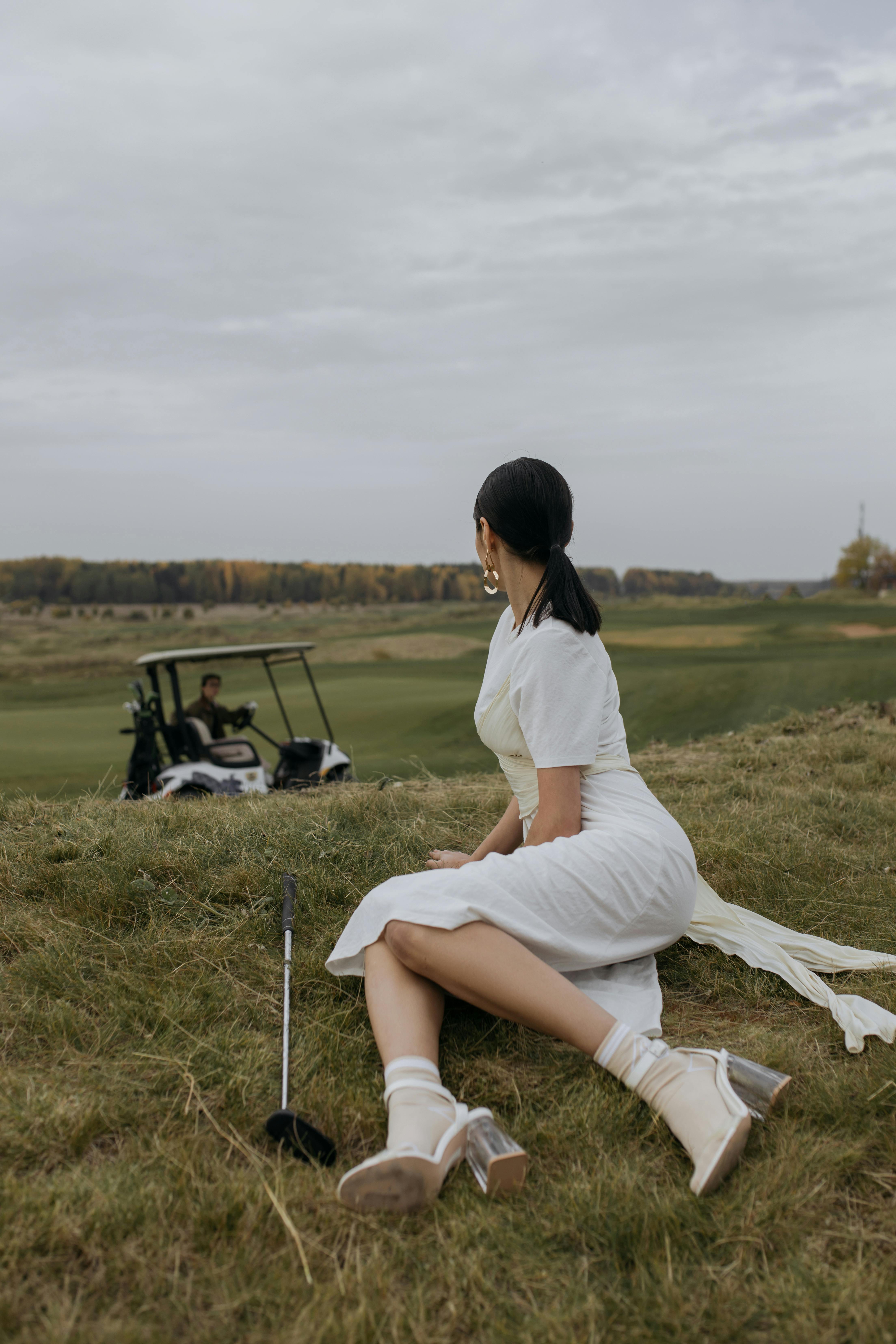 woman in white dress sitting on green grass and waiting for golfer