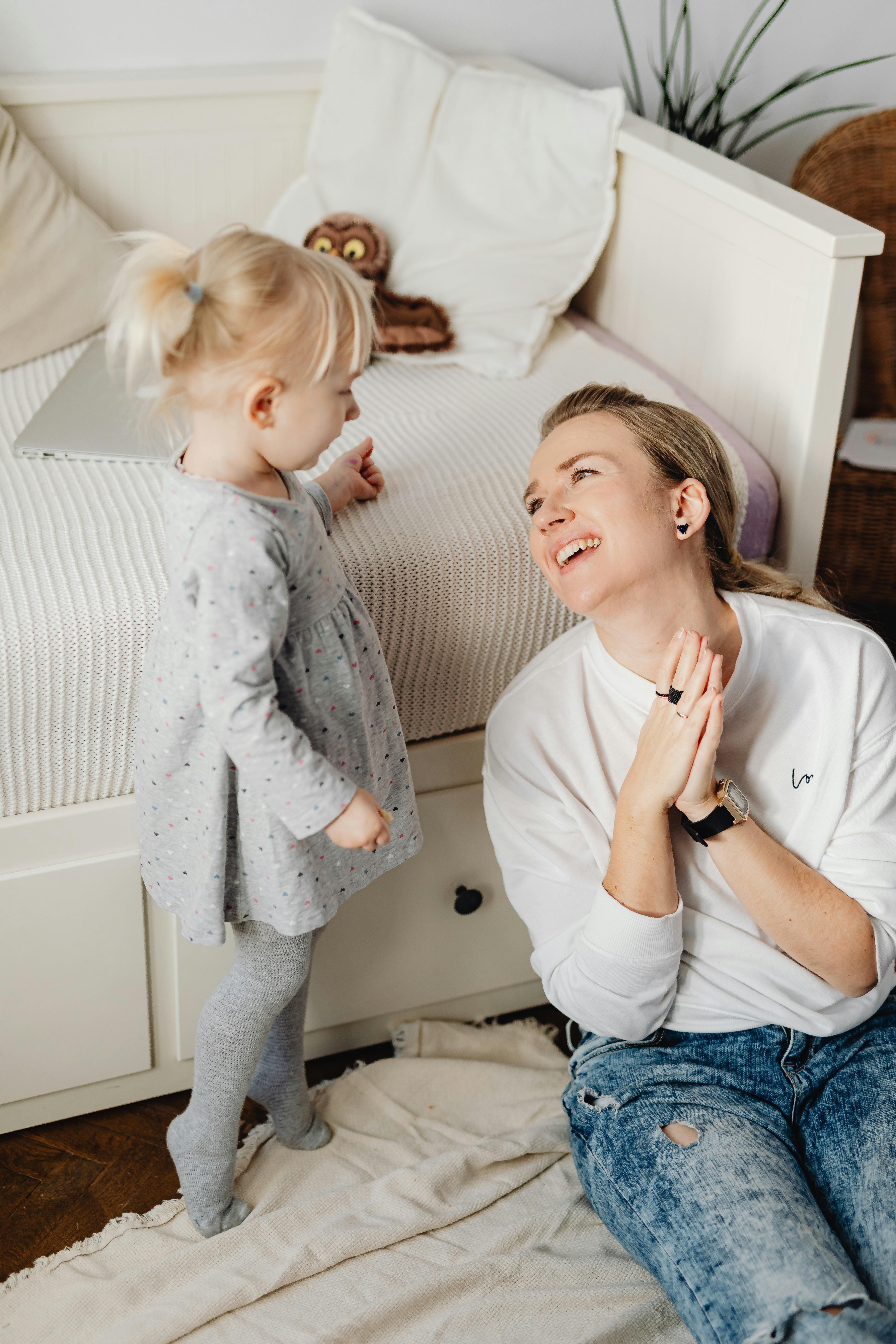 mother sitting next to her little daughter standing near bed