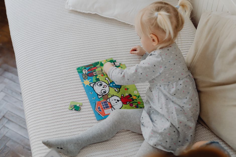 Young girl in a grey dress playing with a colorful puzzle on a couch indoors.