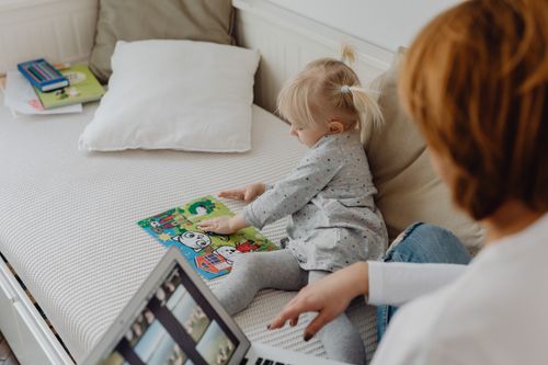 Free A toddler playing with a puzzle next to her mother using a laptop in a cozy home setting. Stock Photo