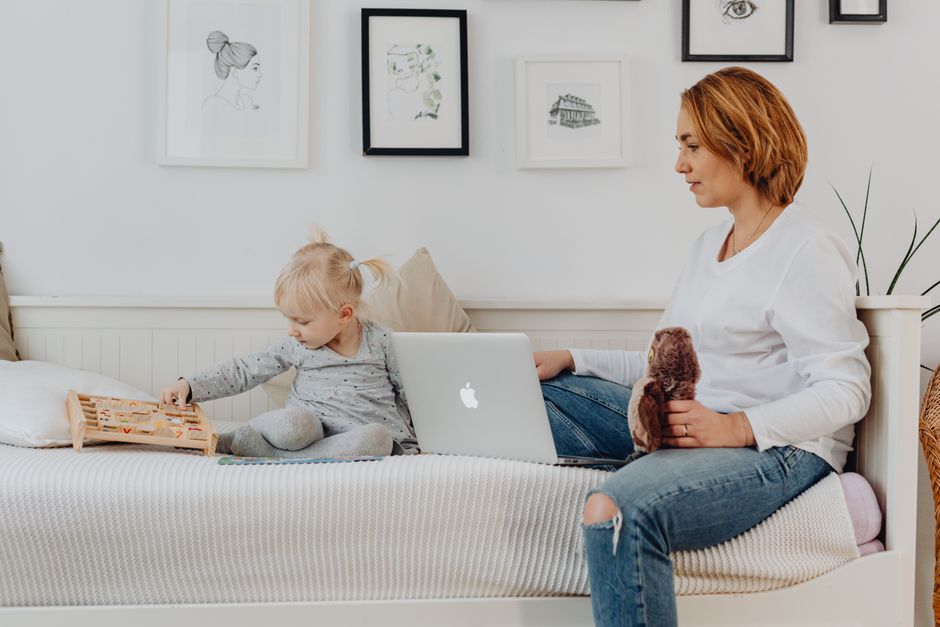 A mother and daughter bonding indoors, sitting on a sofa with a laptop and wooden toy.