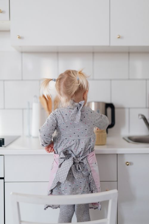 Free Rear view of a toddler girl standing at a kitchen counter, wearing a cute apron. Stock Photo