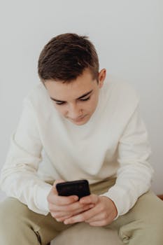 Teenager focused on smartphone, sitting indoors against a white background, symbolizing technology engagement.