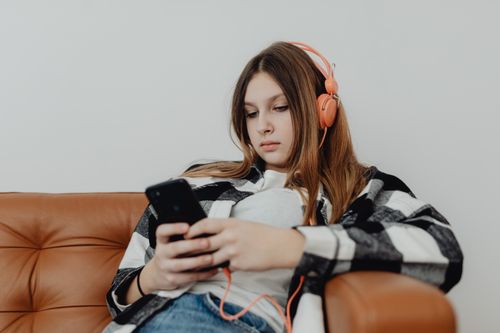 Free Teenage girl sitting on a sofa listening to music on headphones and using a smartphone. Stock Photo