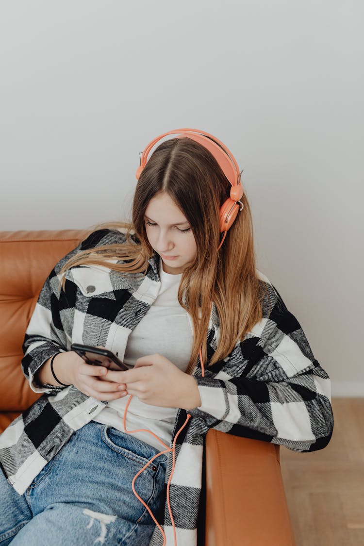 Teenage Girl Sitting On The Sofa With Headphones On And Using Smart Phone