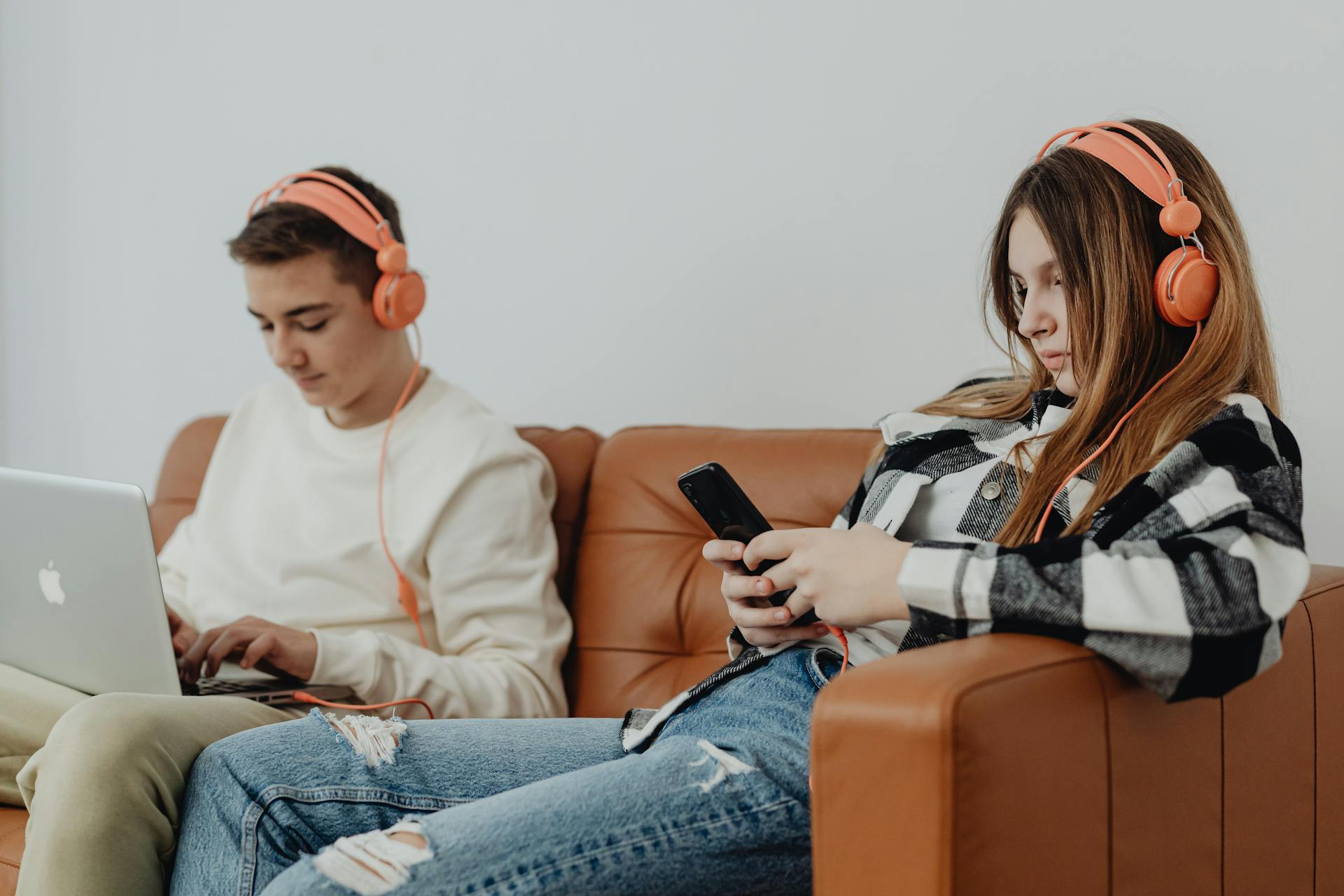 Two teenagers using electronics with headphones on a couch, showcasing modern technology engagement.