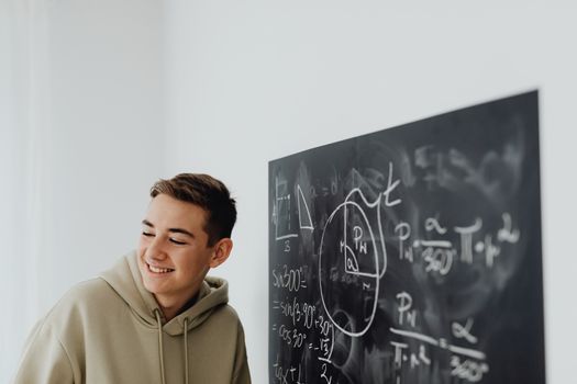 Teen boy in a hoodie smiles while learning algebra on a blackboard indoors.