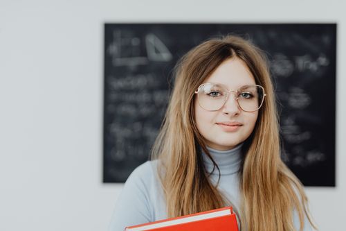 Free Teenage girl with eyeglasses holding books in a classroom with a blackboard background. Stock Photo