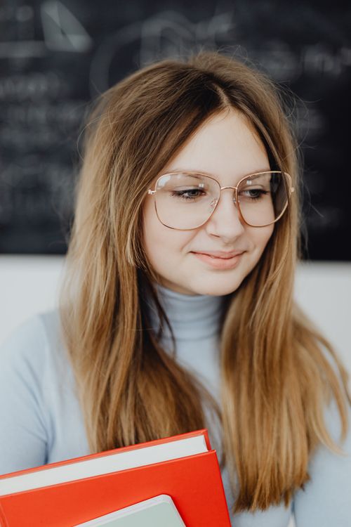Free Portrait of a young woman with glasses holding books in a classroom setting. Stock Photo