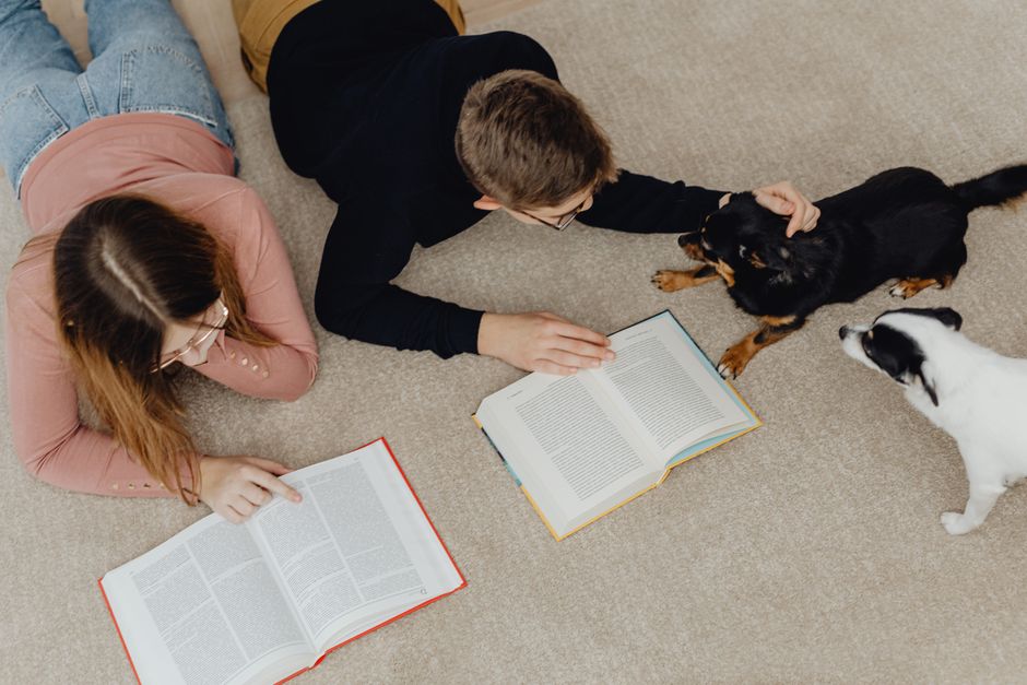Two teenagers reading books on the floor while playing with dogs indoors, emphasizing leisure and companionship.