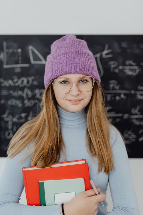 Free Portrait of a young woman in a classroom holding books, wearing glasses and a purple knit hat. Stock Photo