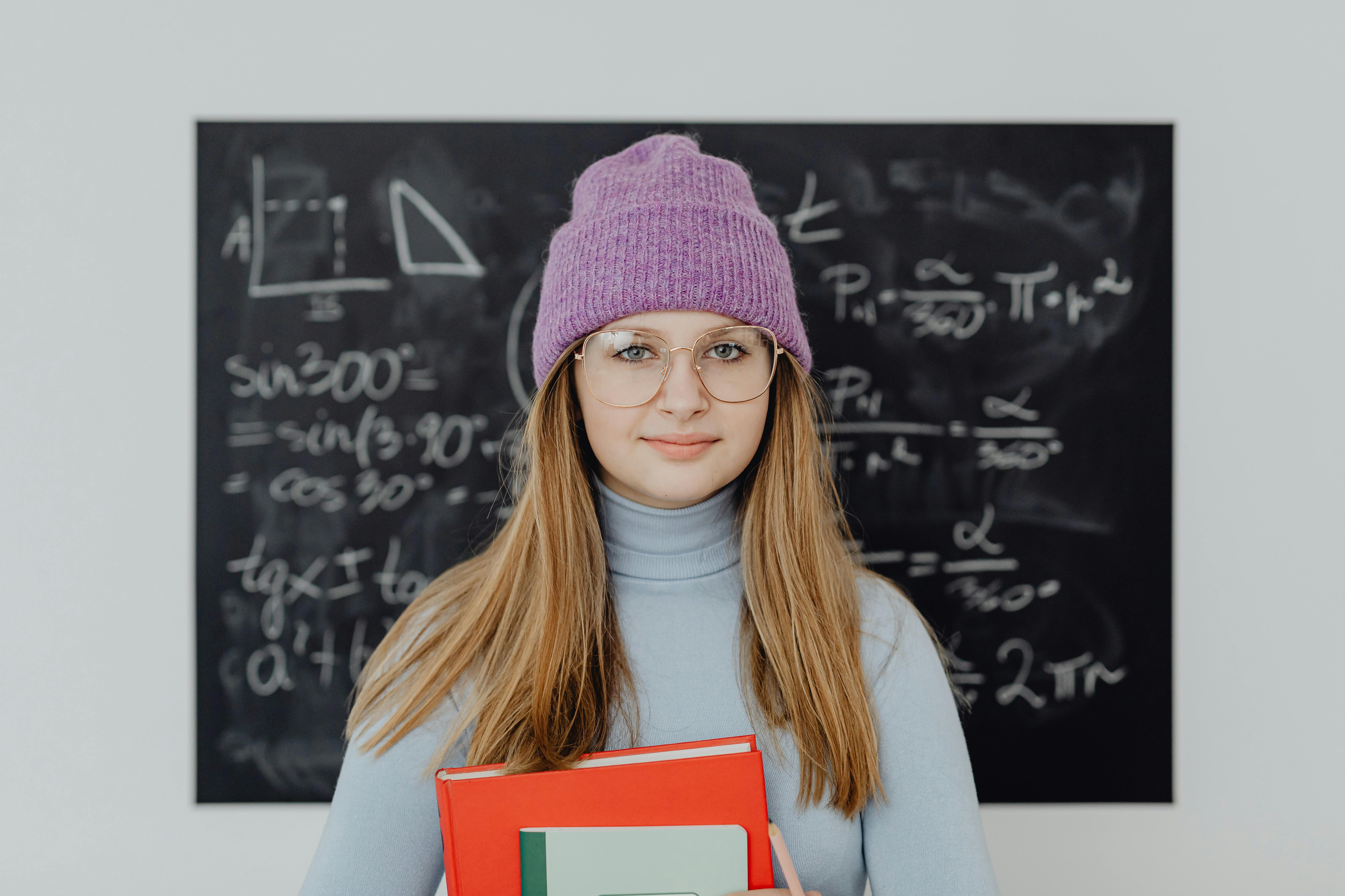 Teenage girl wearing glasses and a beanie, studying math indoors with a blackboard background.