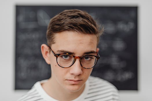 Teenage male student wearing glasses stands in front of a school blackboard.