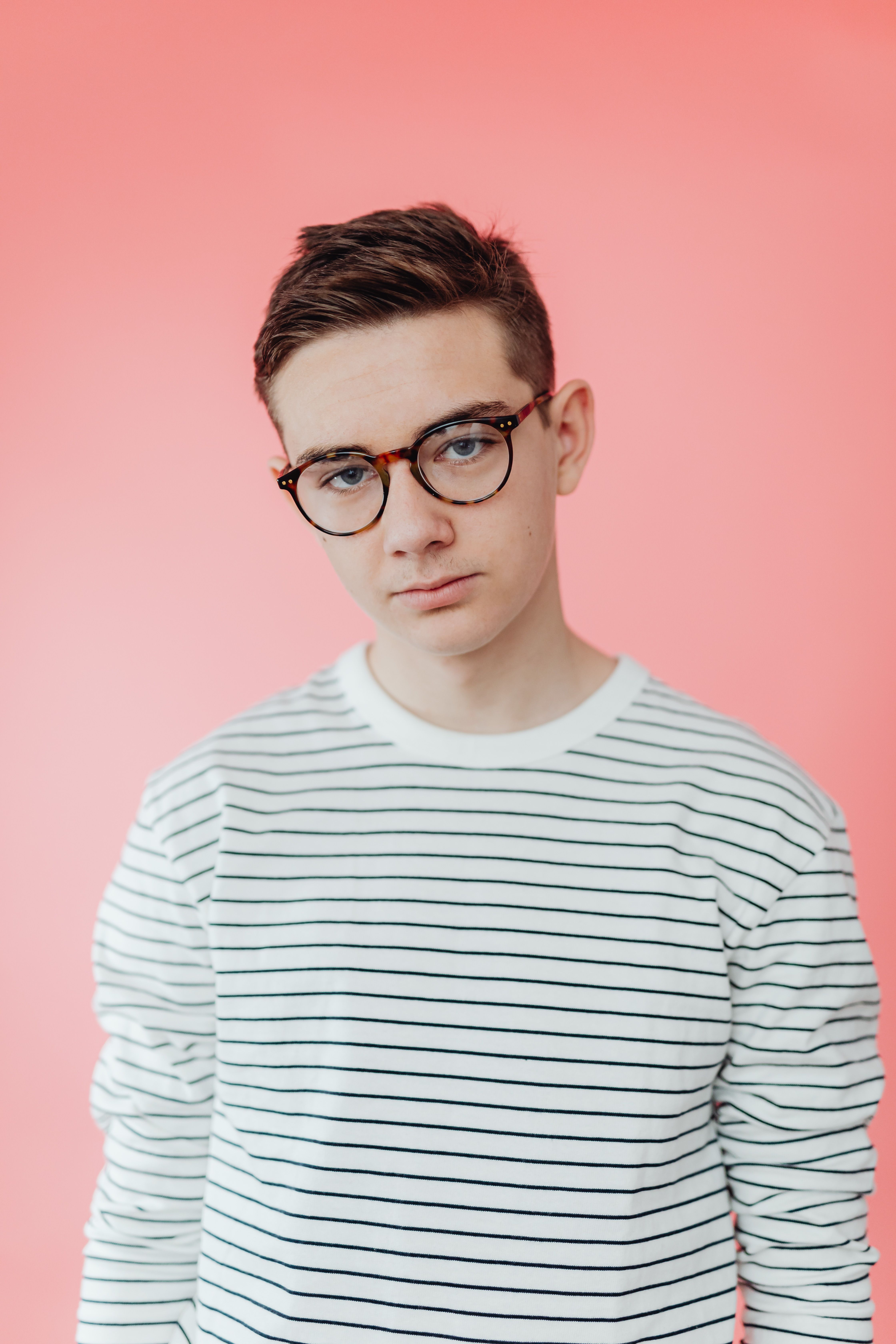 Portrait of a teenage boy wearing glasses and a striped shirt against a pink background.