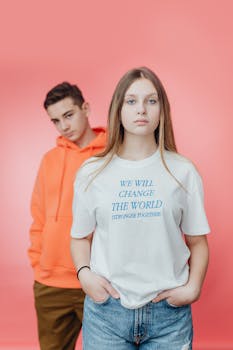 Two young individuals in casual clothing pose confidently against a pink background.