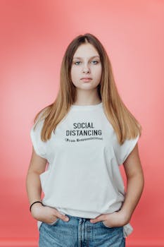 Portrait of a young woman in a white t-shirt with a humorous social distancing message on a pink background.