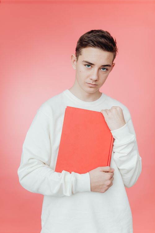 Free Teenage boy in a white sweatshirt holding a book against a pink background in a studio shot. Stock Photo