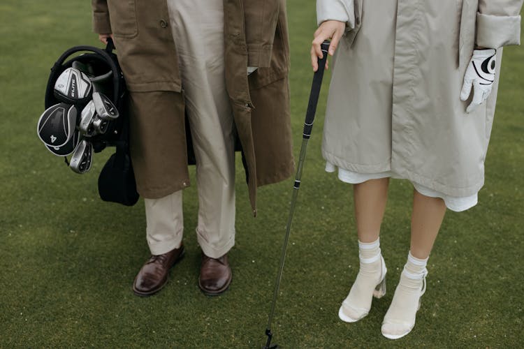 Man Holding A Golf Bag Standing Beside A Woman With Cane On Green Grass 